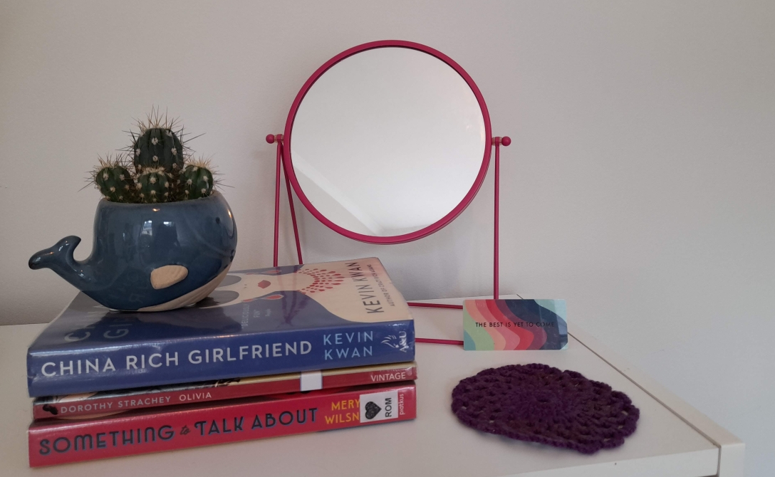 Three books on top of a white shelf. On top of them is a cactus in a pot shaped like a whale. Behind them stands a round pink mirror, and to their right is a purple knitted coaster and a rainbow card with the words 'the best is yet to come' on it